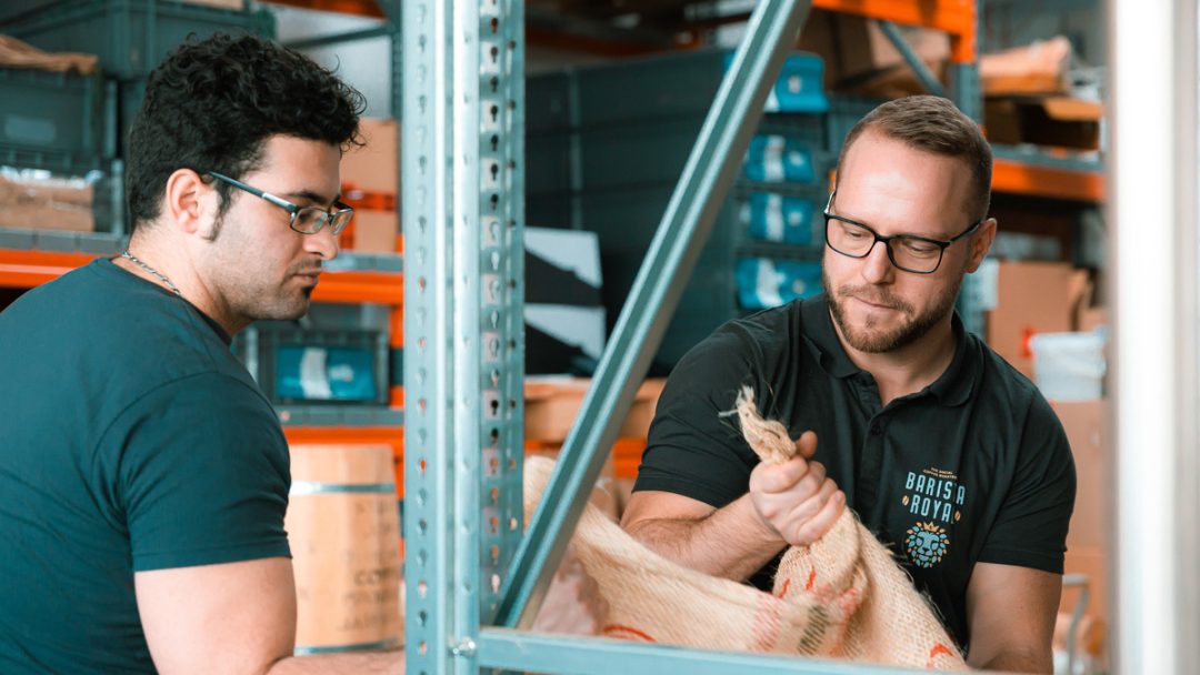 Röster mit Behinderung und Barista Royal Gründer Michael Halbritter heben gemeinsam einen Sack mit Rohkaffeebohnen hoch.