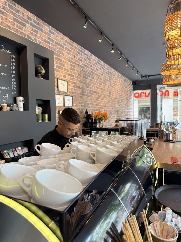 Das Bild zeigt einen Ausschnitt aus dem Café Il Barista Tommy Dee. Man sieht die Oberseite Kaffeemaschine mit weißen Tassen und direkt dahinter Tommy.